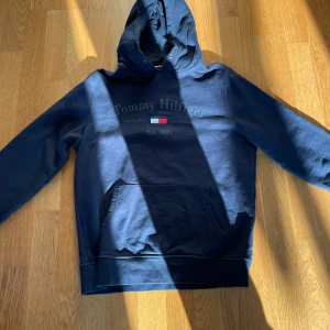 Mörkblå hoodie från Tommy Hilfiger - Säljer en snygg mörkblå hoodie från Tommy Hilfiger med klassisk logga och ficka framtill. Perfekt för en avslappnad stil. Hoodien har långa ärmar och en bekväm passform.