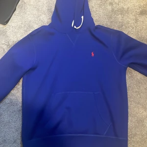 Blå hoodie från Polo Ralph Lauren - Säljer en snygg blå hoodie från Polo Ralph Lauren med det klassiska logotypen broderad i rött på bröstet. Tröjan har en känguruficka och en justerbar huva med snören. Perfekt för en avslappnad stil. Använd 1 gång och är i bra skick.