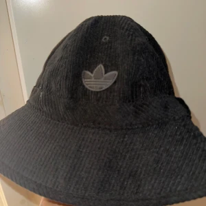 Svart manchesterhatt från Adidas - Manchesterhatt från Adidas med det klassiska logotypen framtill. Hatten har en bred kant och är tillverkad i mjukt manchestertyg. Endast använt en gång.