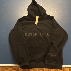 Svart Essentials hoodie - Säljer en helt ny Essentials hoodie med texten 'ESSENTIALS' tryckt på framsidan. Hoodien har en klassisk design med långa ärmar och en rymlig huva. Kom med bud och frågor! Nypris: 2400kr🤗