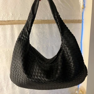 Svart flätad axelväska - Snygg svart axelväska med flätat mönster. Väskan har en praktisk dragkedja och är tillverkad i skinn. Perfekt för att bära med sig det viktigaste med stil.