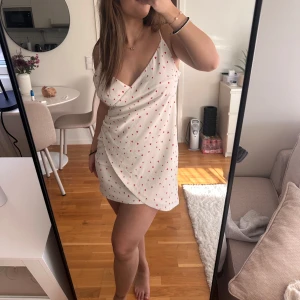 Vit omlottklänning med hjärtan - Söt vit omlottklänning med röda hjärtan. Klänningen har en v-ringad design och är ärmlös, perfekt för sommarens varma dagar. Super söööt! Storlek M ❤️