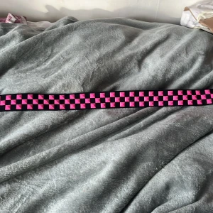 Svart och rosa rutigt y2k bälte - Coolt bälte med svart och rosa rutmönster. Bältet har en rosa spänne som matchar de rosa rutorna. Perfekt för att ge en färgklick till din outfit.