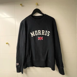 Svart sweatshirt från Morris - Säljer en svart sweatshirt från Morris med vit text och en liten brittisk flagga på framsidan. Tröjan har långa ärmar och en rund halsringning. Perfekt för en avslappnad stil.