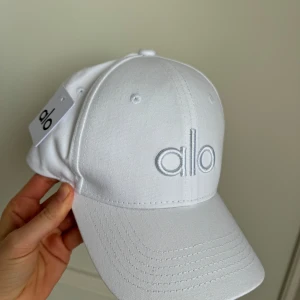 Vit keps från Alo - Snygg vit keps från Alo med broderad logga framtill. Kepsen har en klassisk design med justerbar passform och ventilerande hål för extra komfort. Perfekt för en stilren look.