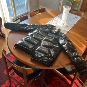 Moncler maya  - Säljer en snygg svart dunjacka från Moncler. Jackan har en glansig finish och är utrustad med dragkedja och en ficka med knappdetalj på ärmen. Perfekt för kyligare väder. Storleken är 3 som det står där men vet inte vad jag ska sätta på Plick så jag bara satta nått. 