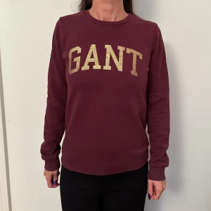 Vinröd tröja från GANT - Säljer en snygg vinröd tröja från GANT med guldglittrig logga på framsidan. Tröjan har långa ärmar och en klassisk rund halsringning. 