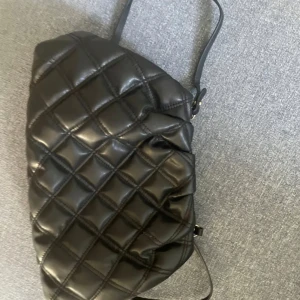Svart quiltad axelväska - Snygg svart quiltad axelväska med dragkedja. Perfekt för att bära dina dagliga nödvändigheter med stil. Den har en ljus insida med en praktisk innerficka för extra förvaring. Säljer då den inte används längre💕💕