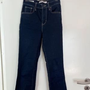 Levi's 725 High Rise Bootcut Jeans - Snygga mörkblå Levi's 725 High Rise Bootcut jeans med hög midja och klassisk femficksdesign. Perfekta för en stilren look med en bootcut passform som ger en tidlös siluett. Tillverkade i slitstarkt denim med kontrasterande sömmar.