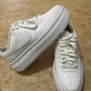 Vita sneakers från Nike - Säljer ett par klassiska vita sneakers från Nike med snygg perforerad design på ovansidan och en rejäl sula. Skorna har snörning och är perfekta för en stilren look. Har använt de 1-2 dagar. Är äkta. 