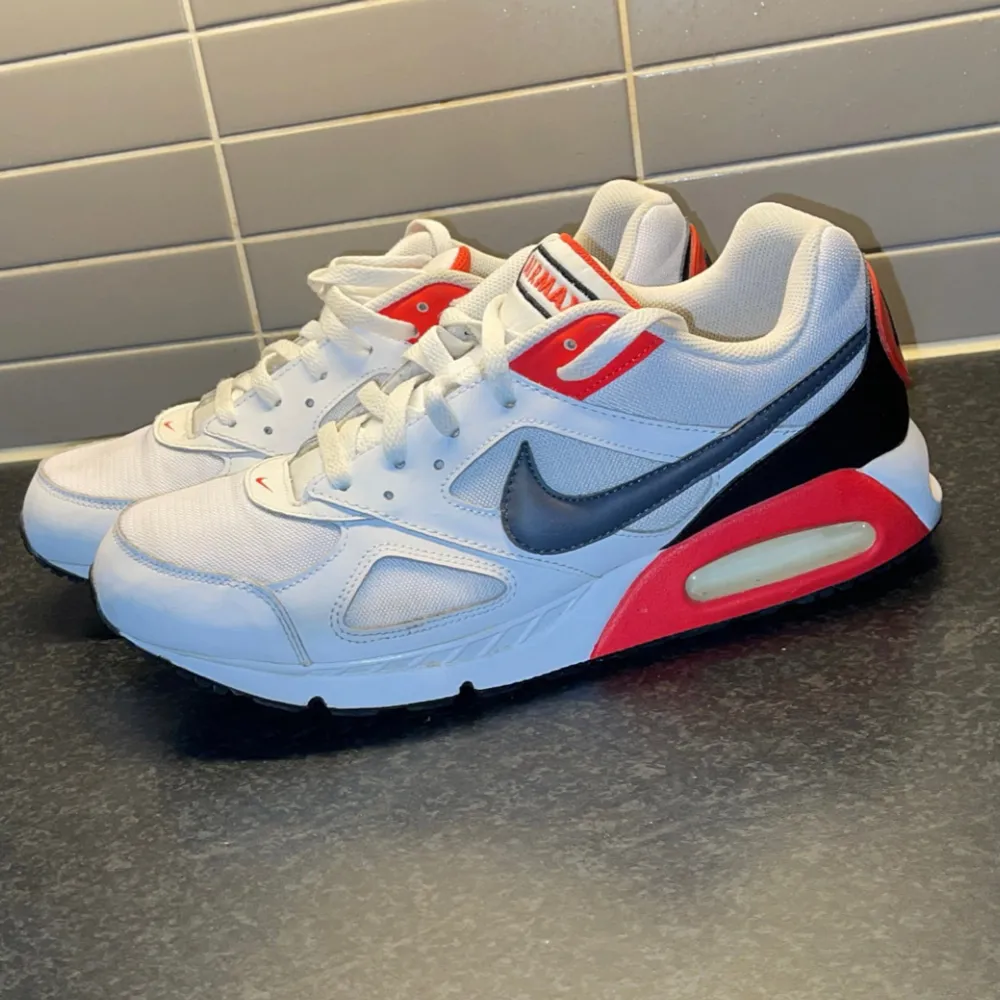 Säljer ett par Nike Air Max 90 sneakers i vitt med mörkblå och röda detaljer. Skorna har en synlig luftkudde i sulan och klassisk snörning. Perfekta för en sportig look! I princip nyskick.. Kengät.
