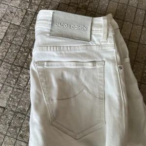 Vita jeans från Jacob Cohën - Snygga vita jeans från Jacob Cohën med klassisk design och knappgylf. Perfekta för en stilren look. Märkesdetaljer på baksidan och framfickor med JC-logga. Lite fläck längst ner på jeansen men syns knappt då man kan vika upp enkelt. Tveka inte att fråga!