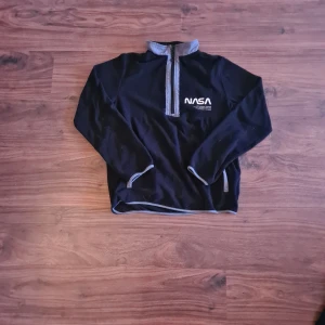 Svart långärmad tröja  - Säljer en svart långärmad tröja från H&M med ett coolt NASA-tryck på bröstet. Tröjan har en dragkedja vid halsen och grå detaljer. 