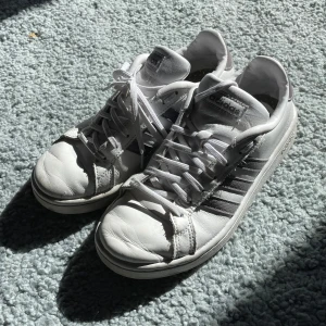 Vita sneakers från Adidas - Säljer ett par klassiska vita sneakers från Adidas med de ikoniska tre ränderna på sidan. Skorna har snörning och en rund tå, perfekt för en avslappnad stil.