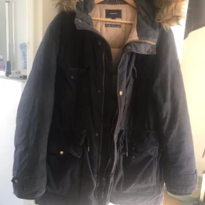 Mörkblå parkajacka från GANT - Säljer en snygg mörkblå parkajacka från GANT med pälskantad huva. Jackan har flera praktiska fickor och stängs med dragkedja och knappar. Perfekt för kyliga dagar! XL