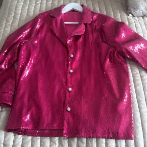 Rosa paljettjacka med matchande byxor - Säljer en glittrig rosa paljettjacka med matchande byxor. Jackan har långa ärmar och knäpps med dekorativa knappar. Byxorna har en knytning i midjan för extra stil. Perfekt för en festlig look! Säljes pga viktnedgång. Passar en storlek 44. Nypris 2700 kr. Från Nadine Merabi. 