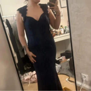 Mörkblå spetsklänning med öppen rygg - Elegant mörkblå långklänning i spets med en vacker öppen rygg och hjärtformad urringning. Klänningen har breda axelband och en figurnära passform som framhäver silhuetten. Perfekt för en festlig kväll! 5 år gammal och använt en gång 