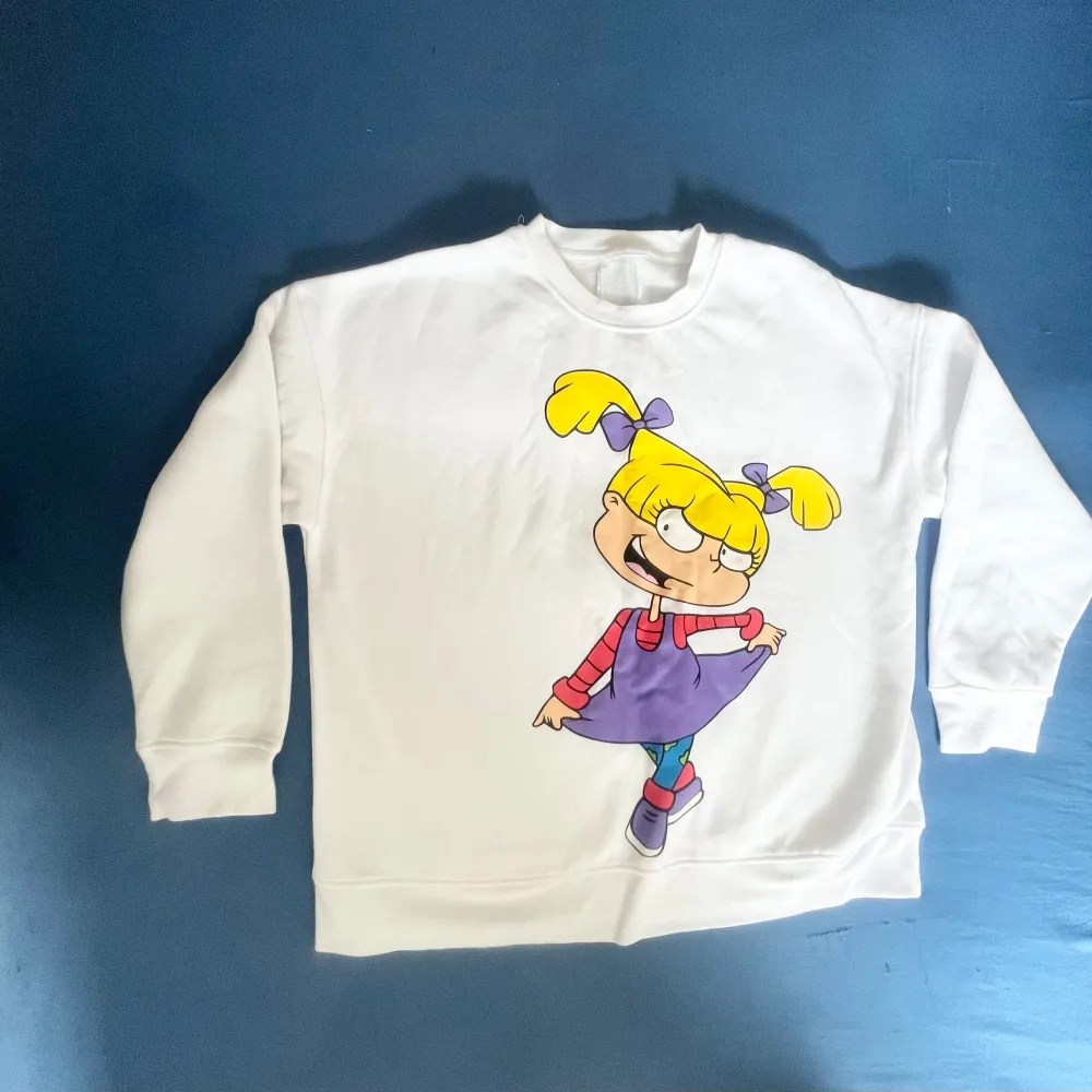 Säljer en vit sweatshirt med en stor färgglad tecknad figur på framsidan. Tröjan har långa ärmar och en rund halsringning. Perfekt för en avslappnad stil med en rolig twist!. Neuletakit & Villapaidat.
