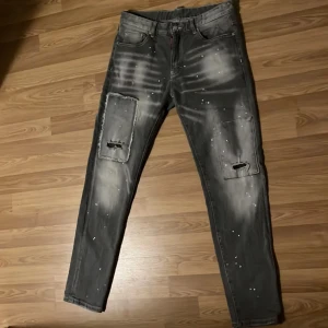 Grå jeans med slitna detaljer - Snygga grå jeans med slitna detaljer och unika mönster. Byxorna har en normal passform och är dekorerade med ljusa fläckar och revor på benen. Perfekta för en avslappnad stil.