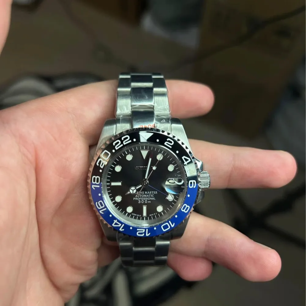 Säljer en automatisk klocka med en stilren design. Klockan har en blå och svart bezel med 24-timmarsmarkeringar och en svart urtavla. Armbandet är i silverfärgat stål och klockan är vattentät upp till 300 meter.. Asusteet.