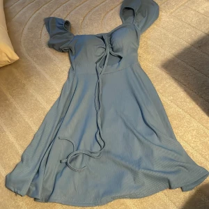 Blå klänning med knytband - Charmig blå klänning med korta ärmar och knytband framtill. Klänningen har en lätt och flödande design som ger en feminin touch.
