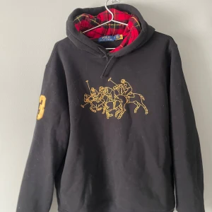 Svart hoodie från Ralph Lauren - Säljer en svart hoodie från Ralph Lauren med ett gult tryck av polospelare på framsidan. Tröjan har en röd och svart rutig insida av huvan och en gul siffra på ärmen. Perfekt för en avslappnad stil.