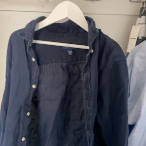 Mörkblå skjorta från GANT - Snygg mörkblå skjorta från SANT med knappar framtill. Perfekt för en stilren look. Skjortan har en klassisk krage och är tillverkad i ett bekvämt material. Perfekt inför sommarn