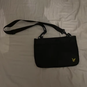 Svart axelväska från lyle scott - Snygg svart axelväska med justerbar rem och dragkedja. Väskan har en diskret logotyp i hörnet och är perfekt för att bära med sig det nödvändigaste. Enkel och stilren design som passar till många tillfällen.