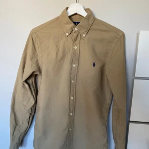 Beige skjorta från Ralph Lauren - Säljer en stilren beige skjorta från Ralph Lauren i slim fit. Skjortan har långa ärmar och knappar framtill samt en broderad logotyp på bröstet. Perfekt för en klassisk look.