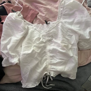 Vit blus med puffärmar - Säljer en söt vit blus med puffiga ärmar och knytning framtill. Blusen har en smockad detalj vid halsen och volangkant vid ärmslut och nederkant. Perfekt för en somrig look!