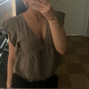 Grå blus med volangärmar - Säljer en söt brun blus med volangärmar och v-ringning. Blusen har en knytning i ryggen och en luftig passform som ger en avslappnad look. Perfekt för en stilren och bekväm outfit.