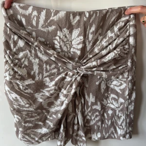 Beige och vit kjol från Gina Tricot - Snygg beige och vit kjol från Gina Tricot med ett blommigt mönster. Kjolen har en knytning framtill som ger en fin detalj och en omlott-look. 