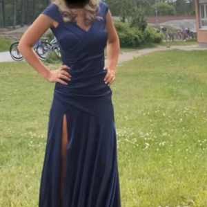 Mörkblå långklänning med slits - Elegant mörkblå långklänning med v-ringning och en stilfull slits framtill. Klänningen har en omlott-design som ger en smickrande passform. Perfekt för festliga tillfällen.