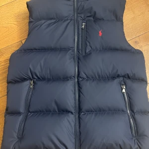 Blå dunväst från Ralph Lauren - Snygg blå dunväst från Ralph Lauren med dragkedja och praktiska fickor. Den har en broderad logga i rött på bröstet. Perfekt för kyligare dagar och även varmare  när du vill hålla stilen.                                                                         Endast använd 2 gånger, ny pris är 2700kr. Passar dig mellan 160-170cm
