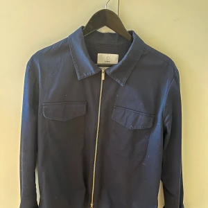 Mörkblå jacka från Bläck - Säljer en stilren mörkblå jacka från Bläck med dragkedja framtill och två bröstfickor. Jackan har en klassisk krage och långa ärmar. Perfekt för en minimalistisk look.