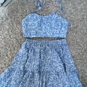 Blå blommig kjol med axelbands topp - Charmig blå och vit blommig axelbands topp och matchande kjol. Perfekt för sommarens varma dagar. Toppen har smala axelband och kjolen är luftig och bekväm. Kom privat för mer bilder