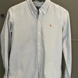 Ljusblå skjorta från Ralph Lauren - Säljer en stilren ljusblå skjorta från Ralph Lauren som e perfekt för sommaren. Skjortan har en broderad logotyp på bröstet och är i storlek M (custom fit-modell) Perfekt för en snygg och avslappnad look. Inga defekter och knappt använd! 😃