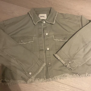 Grön jeansjacka från Monki - Snygg grön jeansjacka från Monki i storlek S. Jackan har en croppad design med fransiga kanter och knappar framtill. Aldrig använd! 