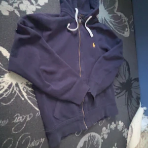 Mörkblå hoodie med dragkedja från Ralph Lauren - Säljer en snygg mörkblå hoodie från Ralph Lauren med dragkedja och huva. Den har en broderad gul logotyp på bröstet och vita snören vid huvan. Perfekt för en avslappnad stil!