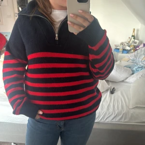 Blå och röd randig tröja med dragkedja - Säljer en svart och röd randig tröja med dragkedja i halsen. Tröjan har långa ärmar och är perfekt för kyligare dagar. Den är både stilren och bekväm, vilket gör den till ett mångsidigt plagg i garderoben. Funkar både på män och kvinnor, inte min stil men skitsnygg på sommaren! Lite nopprig men inga defekter, overzised i passformen 