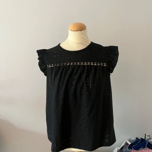 Svart blus med spetsdetaljer - Säljer en elegant svart blus med spetsdetaljer och korta ärmar. Blusen har en lös passform och är perfekt för både vardag och festliga tillfällen.