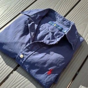 Blå skjorta från Ralph Lauren - Säljer en klassisk blå skjorta från Ralph Lauren med lång ärm och knappar framtill. Skjortan har en broderad röd logotyp på bröstet och är tillverkad i bomull. Perfekt för en stilren look inför sommarn! Den ser lite mörkare ut i verkligheten