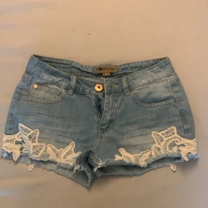 Lågmidjade Jeansshorts med spetsdetaljer - Snygga ljusblå jeansshorts från Lab Industries med fransiga kanter och vita spetsdetaljer på sidorna. Perfekta för en somrig dag, dragkedjan är sönder men annars är dom i bra skick, pris kan diskuteras, skriv om bilder