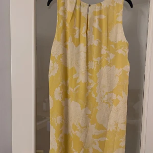 Gul blommig klänning från H&M - Säljer en somrig gul klänning från H&M med ett vackert blommigt mönster i vitt. Klänningen är ärmlös och har en avslappnad passform, perfekt för varma dagar. Den har en rund halsringning och ett lätt och luftigt material.