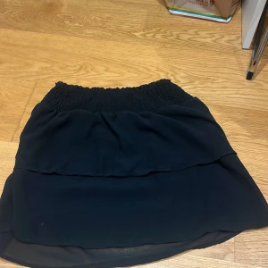 Svart kjol från ONLY - Snygg svart kjol från ONLY i storlek XS. Kjolen har en elastisk midja med smockdetaljer och är tillverkad i ett lätt och flödande material. Perfekt för en stilren look.