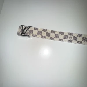 Rutig bälte från  - Snyggt bälte från med klassiskt rutigt mönster i beige och grått. Bältet har ett stilrent -spänne i silverfärg. Perfekt för att ge din outfit en lyxig touch.