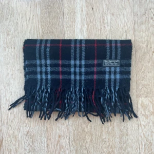 Burberry halsduk - Vintage burberry halsduk i den snygga kombon av svart, grått och de klassiska röda detaljerna. Halsduken är i ett mycket bra skick utan hål, defekter eller fläckar (9/10 skick). Perfekt för att uppgradera din outfit med en snygg accessoar.