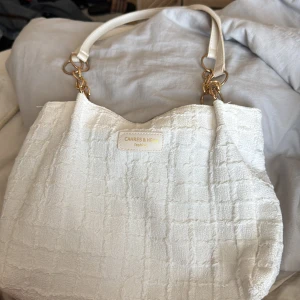 Vit handväska från Cahres & Heirs - Snygg vit handväska från Cahres & Heirs med en elegant textur. Väskan har guldfärgade detaljer och bekväma handtag. Perfekt för att ge en stilren touch till din outfit.