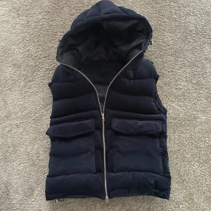 Vendiera Marinblå dunväst med huva - Snygg marinblå dunväst med dragkedja och praktiska fickor. Västen har en mysig huva och är perfekt för kyligare dagar. Den är stilren och enkel att matcha med olika outfits.en defekt på sista bilden,knappen har lossnat men fickan går ändå stängs halvt,har kvar knappen om man vill sy tillbaks den,priset inte hugget i sten!