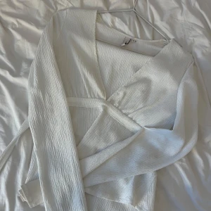 Vit omlottopp - Säljer en stilren vit omlottopp med långärmade ärmar. Toppen har en ribbad struktur och ett knytband i midjan för en elegant look. Perfekt för att skapa en chic outfit. 💫